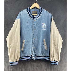 Può includere: Una giacca varsity in denim blu con maniche lunghe beige, caratterizzata da chiusura frontale con bottoni, colletto e polsini a coste a righe e un logo Intel Inside ricamato sul petto. La giacca include due tasche anteriori con finiture beige.