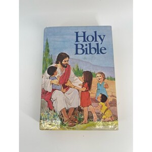 Puede incluir: Una Santa Biblia de tapa dura con una cubierta azul claro que presenta una ilustración a color de una figura con túnicas blancas y rojas sentada al aire libre con cuatro niños. El texto Holy Bible está impreso en azul oscuro arriba.