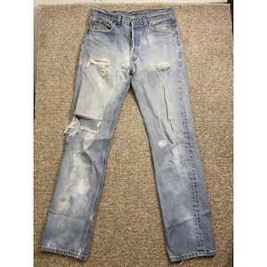 Puede incluir: Vaqueros de mezclilla azul claro con detalles desgastados, incluyendo roturas en las rodillas y parches descoloridos. Los vaqueros tienen un diseño clásico de cinco bolsillos y bragueta con botones.