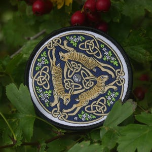 May include: A circular embroidered patch featuring three tan hares arranged in a triangular pattern against a deep blue background. The design includes intricate gold-coloured Celtic knotwork, small white flowers with green leaves, and a black border.