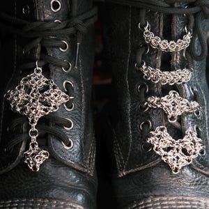 Puede incluir: Primer plano de botas de cuero negro adornadas con dijes de encaje de cota de malla plateada. Los dijes presentan intrincados diseños geométricos, agregando un toque gótico único al calzado. Las botas tienen ojales metálicos y cordones negros.