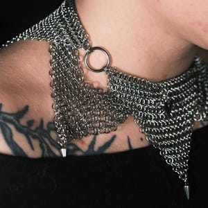 May include: A silver chainmail collar necklace with a large ring clasp and pointed accents. The necklace is made of small, interlocking metal rings, creating a textured, metallic appearance. The design is a unique piece of jewelry.