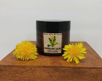 Puede incluir: Un frasco de Dandelion Salve de Death Moth Apothecary se encuentra sobre una caja de madera, flanqueado por dos dientes de león amarillos. El frasco tiene una tapa negra y una etiqueta con el nombre del producto y los ingredientes.