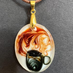 Puede incluir: Collar con colgante ovalado con un diseño temático de café. El colgante presenta una ilustración de una taza de café en negro y verde, sobre un fondo blanco con remolinos marrones y naranjas. El colgante está suspendido de un cordón marrón.
