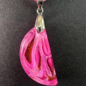 May include: A pink resin pendant with a silver-colored bail hangs from a pink cord. The pendant has a semi-circular shape with swirling patterns of pink, white, and brown. The necklace is displayed against a dark background.
