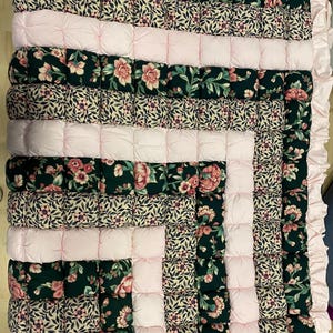 Può includere: Una trapunta fatta a mano con un motivo floreale in verde scuro e rosa. La trapunta presenta file alternate di stampa floreale e quadrati rosa pieni, con un bordo rosa a balze. Un tessuto per la casa accogliente e decorativo.