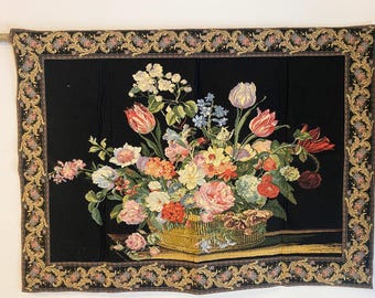 Op de afbeelding: Bloementapijt met een mand vol kleurrijke bloemen, waaronder tulpen en rozen, tegen een zwarte achtergrond. Het tapijt heeft een sierlijke rand en is gemonteerd op een houten roede.