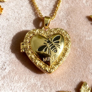 May include: A gold heart-shaped locket necklace featuring a bee design. The locket has a decorative border and the word "MINE" is printed on the front. The necklace is suspended from a gold chain.