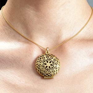 May include: A gold-colored necklace with a round pendant. The pendant features an openwork design with intricate floral patterns. The necklace is worn around the neck, and the chain is delicate and thin.