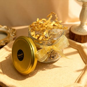 May include: A clear glass jar filled with yellow crinkle paper, topped with a gold lid featuring a black circular sticker that reads Handmade With Love. A gold sheer ribbon is tied around the jar neck, accented with a small gold bow charm.