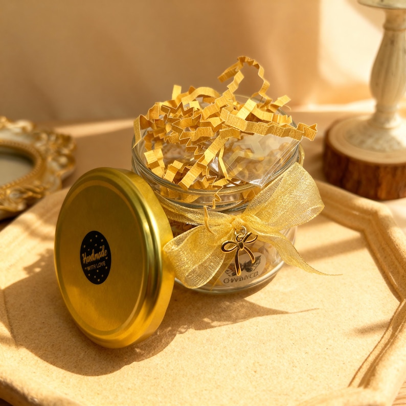 May include: A clear glass jar filled with yellow crinkle paper, topped with a gold lid featuring a black circular sticker that reads Handmade With Love. A gold sheer ribbon is tied around the jar neck, accented with a small gold bow charm.