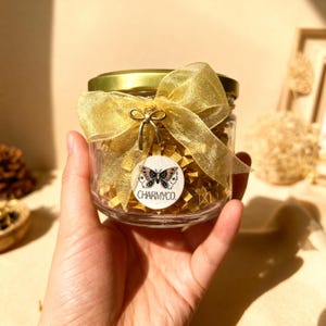 May include: A clear glass jar with a gold lid, adorned with a shimmering gold organza ribbon tied in a bow. A small gold bow charm and a circular white tag featuring a moth illustration and the text CHARMYCO. are attached to the ribbon.