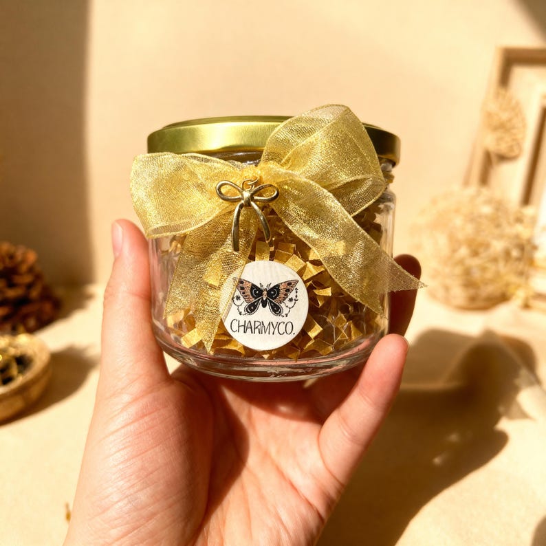 May include: A clear glass jar with a gold lid, adorned with a shimmering gold organza ribbon tied in a bow. A small gold bow charm and a circular white tag featuring a moth illustration and the text CHARMYCO. are attached to the ribbon.