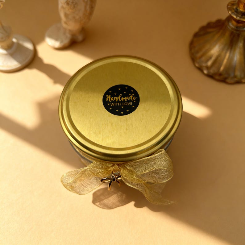 May include: A round jar with a metallic gold lid featuring a circular black sticker that reads Handmade WITH LOVE in gold lettering. A sheer gold ribbon is tied around the neck of the jar, accented with a small metallic charm.