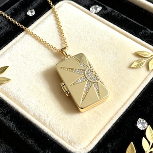 May include: A gold-toned rectangular locket pendant on a matching chain. The front of the locket features a sunburst design with a semi-circular sun motif at the top right, inlaid with small clear crystals that catch the light.