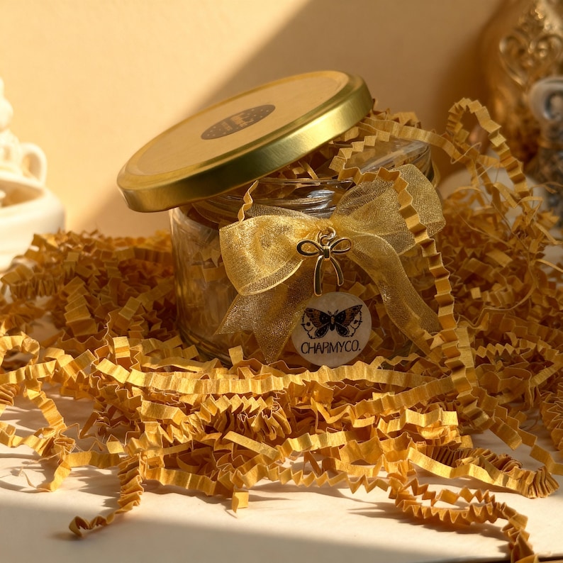 May include: A clear glass jar with a gold lid, adorned with a gold organza ribbon bow and a circular white tag featuring a black butterfly illustration and the text CHARMYCO. The jar is nestled in a bed of crinkled gold paper filler strips.