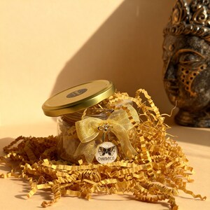 May include: A clear glass jar with a gold lid, filled with shredded yellow paper. A gold organza ribbon is tied around the jar, featuring a circular white tag with a black butterfly illustration and the text CHAPMYCO.