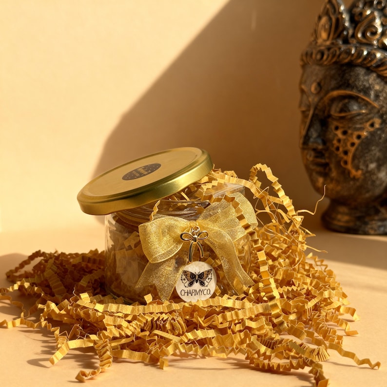 May include: A clear glass jar with a gold lid, filled with shredded yellow paper. A gold organza ribbon is tied around the jar, featuring a circular white tag with a black butterfly illustration and the text CHAPMYCO.