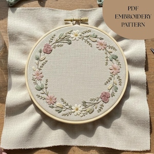 以下が含まれることがあります： ピンクと白の花と緑の葉のリースが特徴の刺繍フープ。刺繍はベージュの生地に施されており、「PDF EMBROIDERY PATTERN」の文字が茶色の円で表示されています。