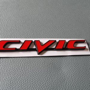 May include: A car emblem featuring the word CIVIC in bold, stylized capital letters. The lettering has a bright red finish with a black border, mounted on a white rectangular adhesive backing strip for application to a vehicle exterior.