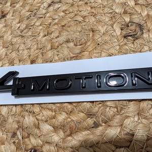 Op de afbeelding: Een zwart embleem met de tekst "4 MOTION" in een glanzende afwerking. Het rechthoekige embleem ligt op een witte strook tegen een geweven achtergrond. Het cijfer "4" staat links.