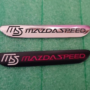 Op de afbeelding: Twee MazdaSpeed-emblemen. Eén is zilverkleurig met zwarte en rode tekst, de andere is zwart met witte en rode tekst. De tekst luidt "MS MAZDASPEED". De emblemen liggen op een groene ondergrond.