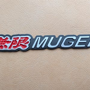 Può includere: Un emblema Mugen nero e argento con caratteri giapponesi rossi. L'emblema ha un design in rilievo ed è probabilmente per un veicolo. Il testo è delineato in argento e i caratteri giapponesi sono rossi.