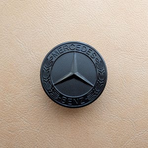 Puede incluir: Un emblema negro de Mercedes-Benz con una estrella de tres puntas en el centro. El emblema circular tiene las palabras "MERCEDES" y "BENZ". El emblema está sobre un fondo beige.