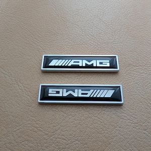 Puede incluir: Dos emblemas AMG negros y plateados. Cada emblema rectangular tiene el logotipo AMG en blanco. El fondo es una superficie texturizada de color marrón claro.