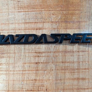 Könnte beinhalten: Ein mattschwarzes Emblem mit dem Wort MAZDASPEED in einer fetten, serifenlosen Schriftart. Die Buchstaben sind in einem Stück verbunden und zeigen ein klares, modernes Design mit scharfen Kanten und einer flachen Oberfläche für den Außenbereich.