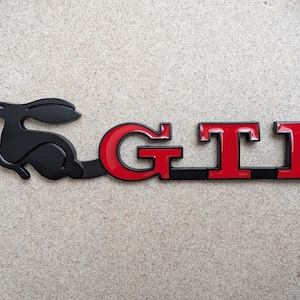 Peut inclure: Emblème GTI noir et rouge avec un motif de lapin. Les lettres GTI sont rouges avec des contours noirs, et le lapin est noir. Conçu pour une utilisation automobile.