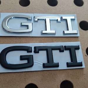 Peut inclure: Deux emblèmes GTI, un chromé et un noir, sont présentés sur un support gris clair. Les lettres sont en gras et carrées, conçues pour l'automobile. Le fond est un panneau perforé marron.