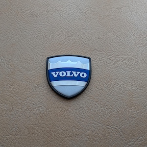 May include: A Volvo emblem with a shield shape. The emblem features a silver and blue design with the word "VOLVO" in bold blue letters. The emblem has a black border and is set against a brown textured background.