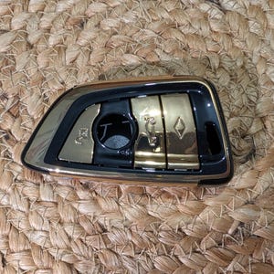 May include: A car key fob with a gold and black design. The fob has several buttons and a keyhole. The key fob is on a woven, tan surface.