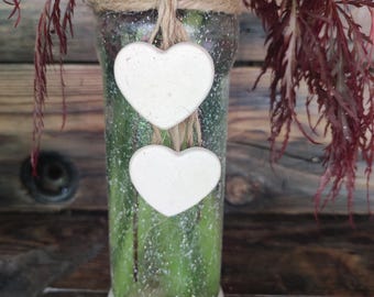 May include: A clear glass vase with a white base and two heart-shaped ornaments hanging from twine. The vase contains green stems and is decorated with glitter. The top of the vase is wrapped with twine, and the background is a wooden surface.