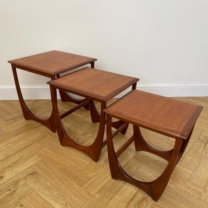 May include: A set of three teak nesting tables. Each table has a square top and curved legs. The wood has a rich, reddish-brown colour. The largest table is approximately 61 cm wide.