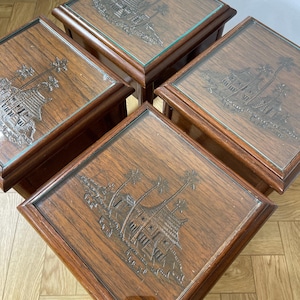 May include: Four square wooden side tables with glass tops. The dark brown tables feature a carved design of a building and palm trees. The tables are arranged in a diamond shape, showcasing the intricate details of the wood and glass.