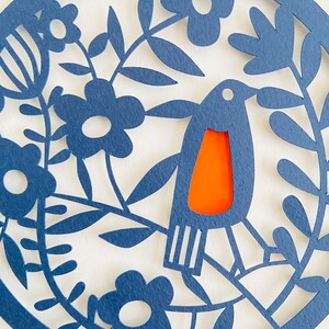 May include: A blue paper cut-out design of a bird with an orange breast, surrounded by flowers and leaves.