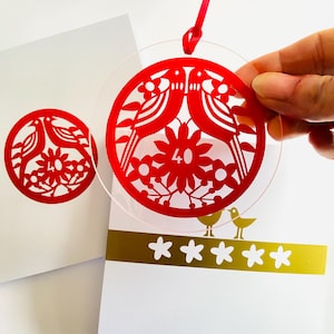 Może przedstawiać: Czerwona, okrągła ozdoba z motywem ptaków i kwiatów oraz liczbą 40. Ozdoba trzymana jest w dłoni, z dołączoną czerwoną wstążką. Pasujący wzór jest nadrukowany na białej karcie. Widoczny jest również złoty pasek z wycięciami w kształcie gwiazd.