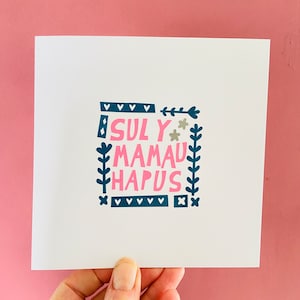 May include: A white square card with the words "SULY MAMAU HAPUS" in pink, teal, and gray. The text is surrounded by teal floral and geometric designs. The card is held against a pink background.