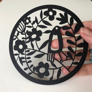 May include: Black paper cut-out design of a bird with flowers and leaves. The design is intricate and detailed, with a delicate silhouette.
