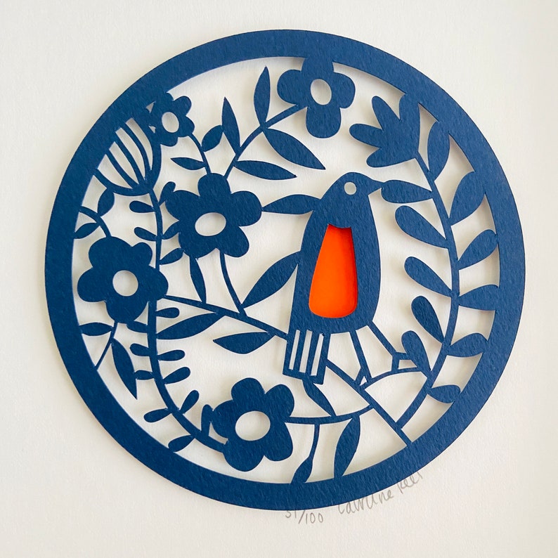 May include: A blue paper cut-out design of a bird with an orange breast, surrounded by flowers and leaves. The design is on a white background and includes the text "51/100  Caroline Rice".