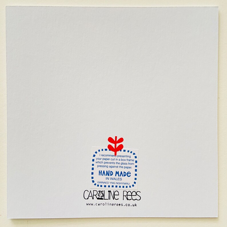 May include: A white card with a blue and red sticker that says "I recommend presenting your paper cut in a box frame which prevents the glass from pressing against the paper. HAND MADE IN WALES GWNAED YNG NGHYMRU CAROLINE REES www.carolinerees.co.uk"