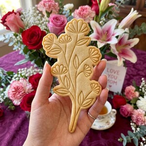 May include: A light brown, flower-shaped cookie with detailed floral designs is held in front of a bouquet of flowers. The cookie has a stem and several flower clusters. A card with the text "LOVE YOU MOM" is visible.