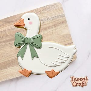 May include: A white goose-shaped cookie with a green bow, orange beak and feet, and pink cheeks. The cookie is decorated with white icing and sits on a wooden cutting board. The cookie has the words "Invent Craft" in the bottom right corner.