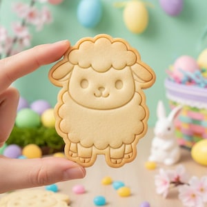 May include: A light brown, sheep-shaped cookie cutter held by a hand. The cookie cutter has a detailed outline of a sheep with a fluffy body and a smiling face. Easter-themed decorations are in the background, including eggs and a basket.