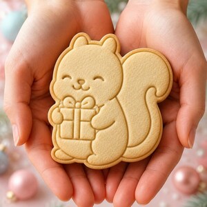 May include: A light beige, flat, squirrel-shaped cookie with a smooth surface. The cookie features an embossed design of a squirrel with a smiling face, holding a small square gift box tied with a ribbon in its front paws, and a large bushy tail.