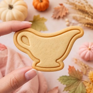 May include: A light tan, smooth-textured sugar cookie shaped like a gravy boat. The cookie features a raised, embossed outline of the vessel's handle, rim, and base, creating a distinct silhouette against the flat, solid center of the baked dough.