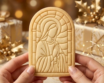 Könnte beinhalten: Ein hellbeiger, bogenförmiger Keks mit einem geprägten Motiv einer Figur mit gefalteten Händen, eingerahmt von einem stilisierten Fensterbogen. Der Keks hat eine glatte, gebackene Oberfläche mit definierten Linien für das detaillierte Bild.