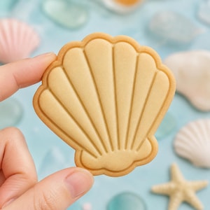 May include: A light tan, shell-shaped cookie with a smooth surface and embossed vertical ridges radiating from the base. The cookie features a scalloped edge and a distinct outline, presenting a clean, uniform appearance in a simple, flat design.